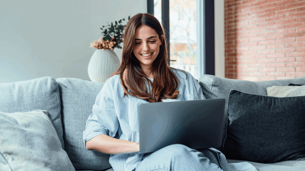 A smiling woman is sitting on a couch with a laptop on her lap.