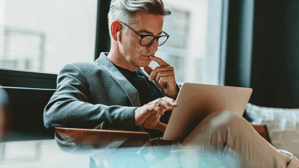 A man in glasses is sitting in a chair and reading from a laptop.
