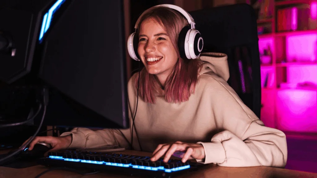 A smiling young woman gamer in headphones is sitting at a computer.