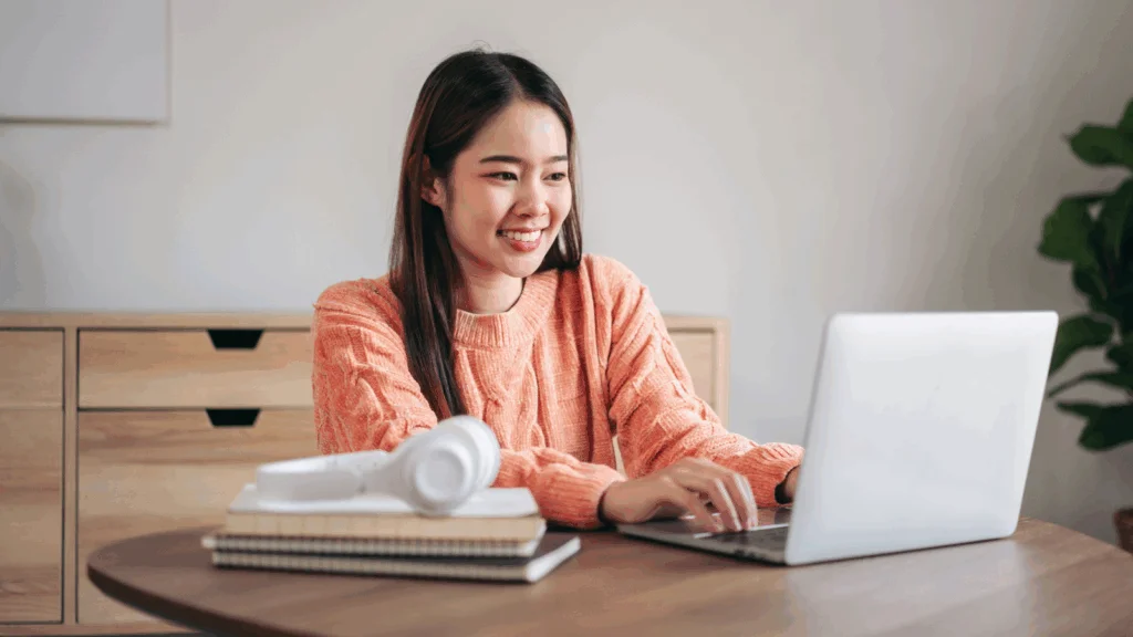 A smiling young woman is sitting at a desk and using a laptop to find out how nonprofits make money.