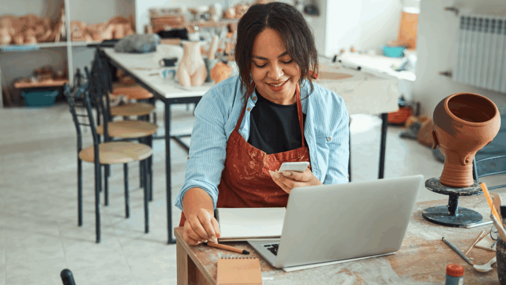 A woman artist is sitting in front of a laptop with a mobile phone in her hand.