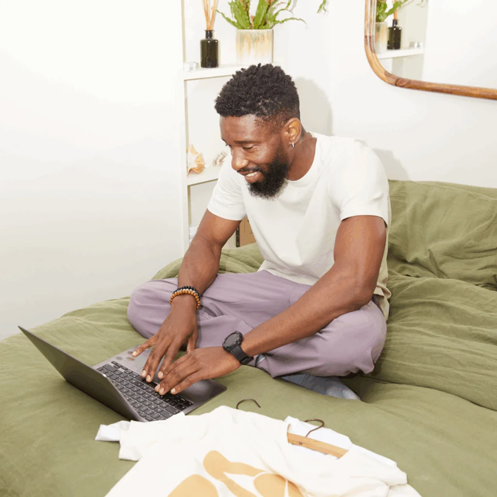 A smiling man is sitting on a bed, using a laptop, with a custom t-shirt laying near the laptop.