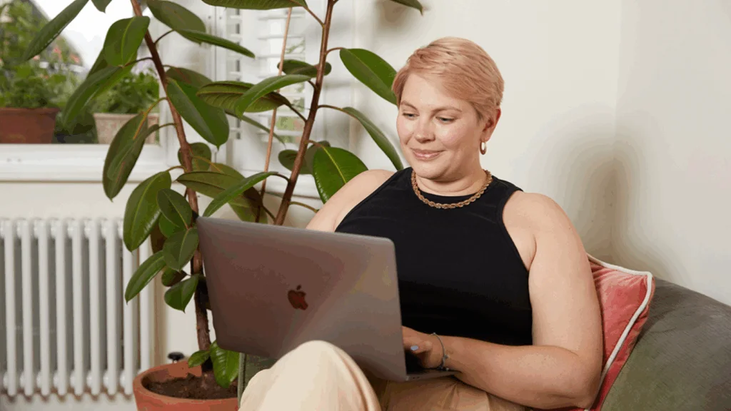 A young woman is sitting in a soft chair and using a laptop.