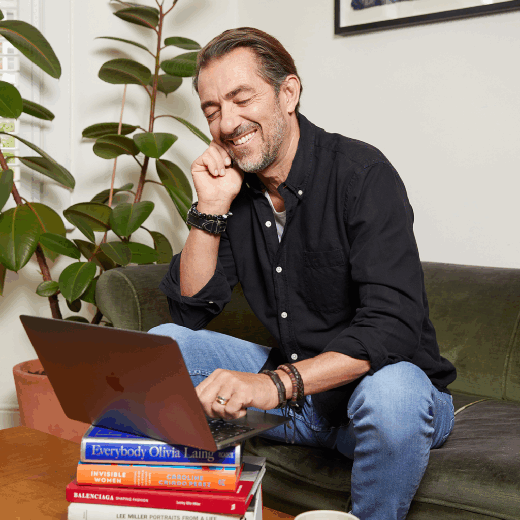 A man is sitting on a couch and using a laptop that’s standing on a stack of books.