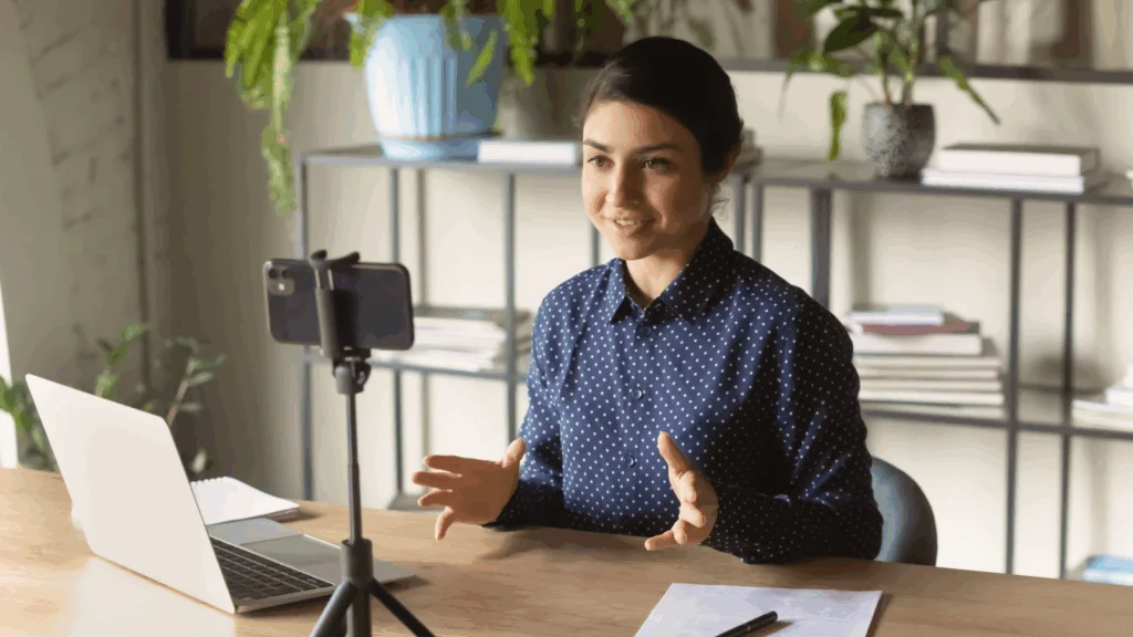 A woman is sitting at a desk with a laptop and recording herself using a mobile phone.