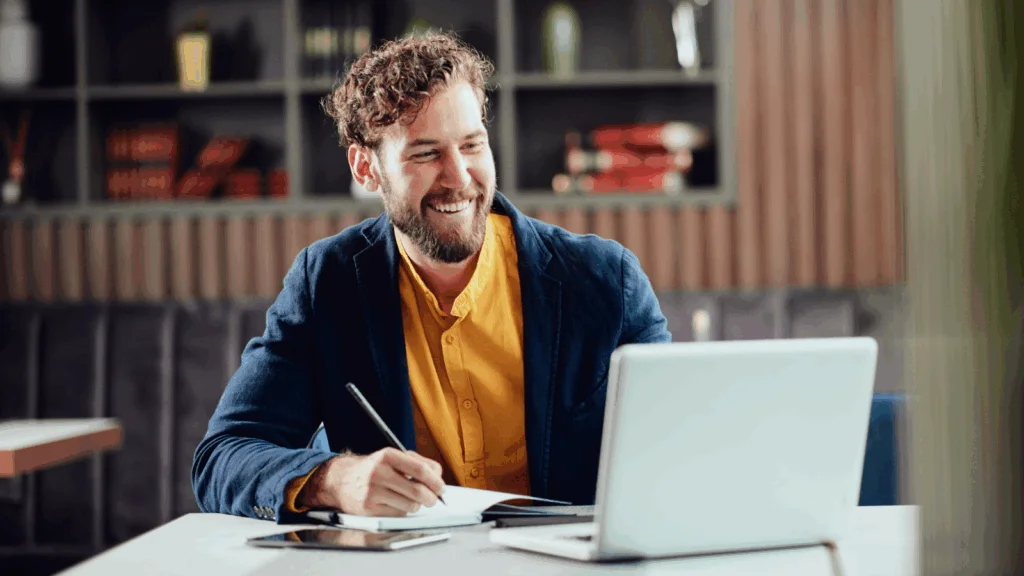 A smiling man is using a laptop and making notes on how to be your own boss.