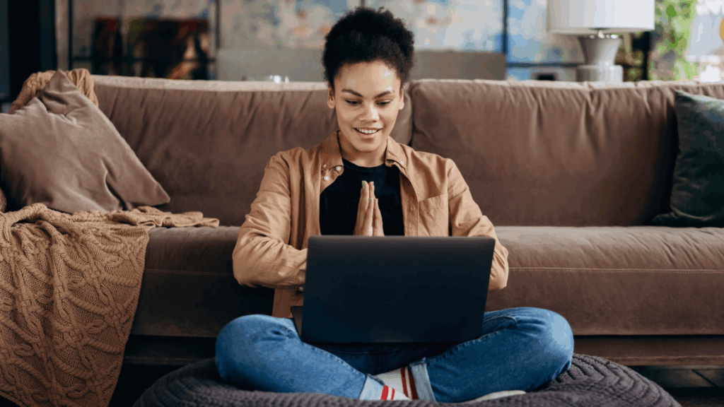 A woman is sitting on the floor with a laptop on her lap, and a couch behind her.