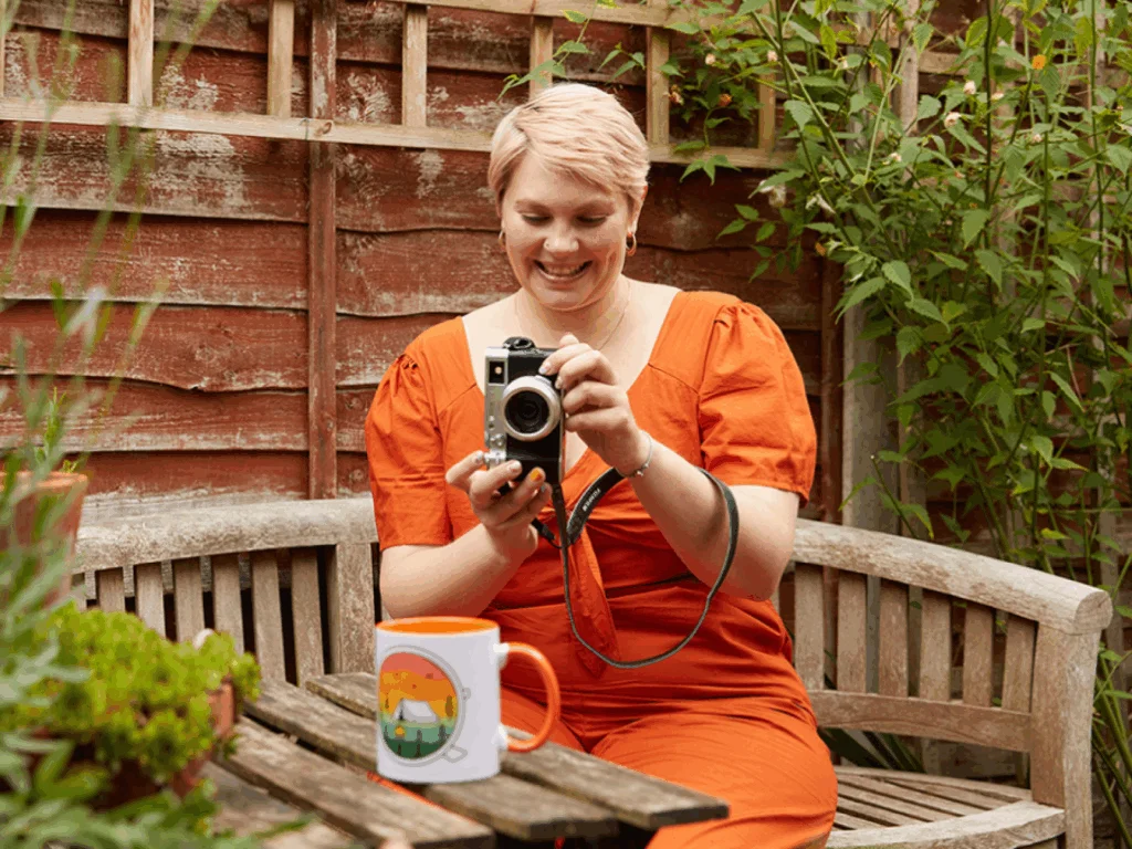 A smiling young woman in an orange dress is taking a picture of a custom mug.