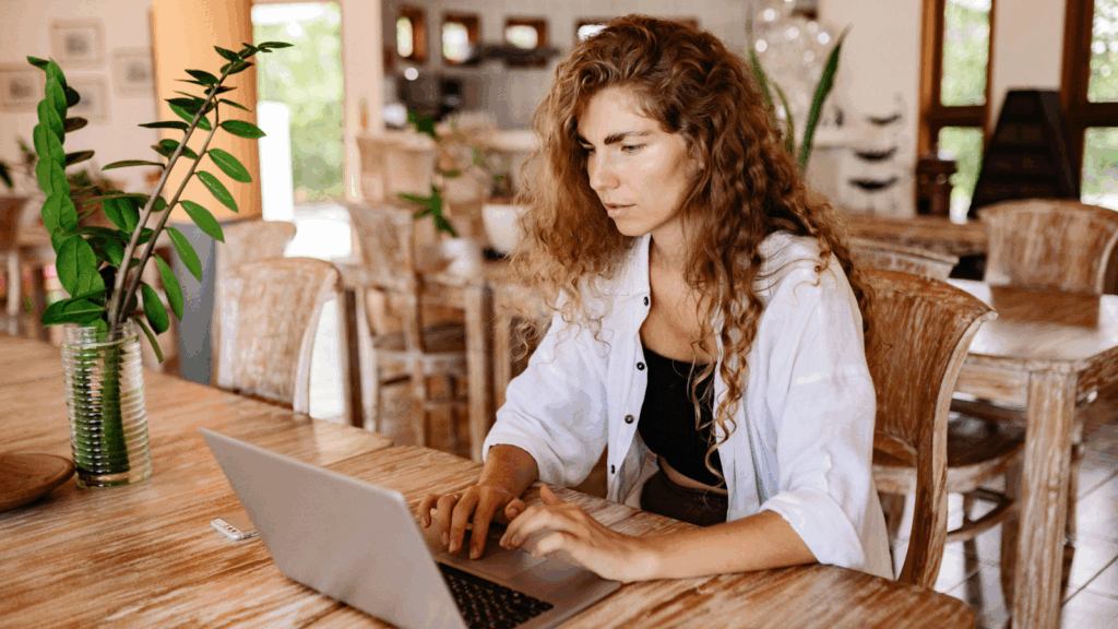 A woman seated at a table, focused on her laptop, with a calm expression in a well-lit room.