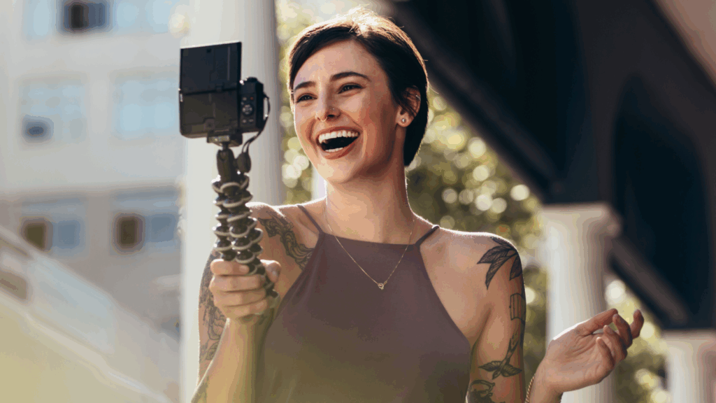 A smiling woman is recording herself using a digital camera on a tripod.