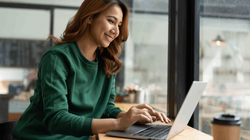 A woman sits at a table, focused on her laptop, with a calm expression in a well-lit room.