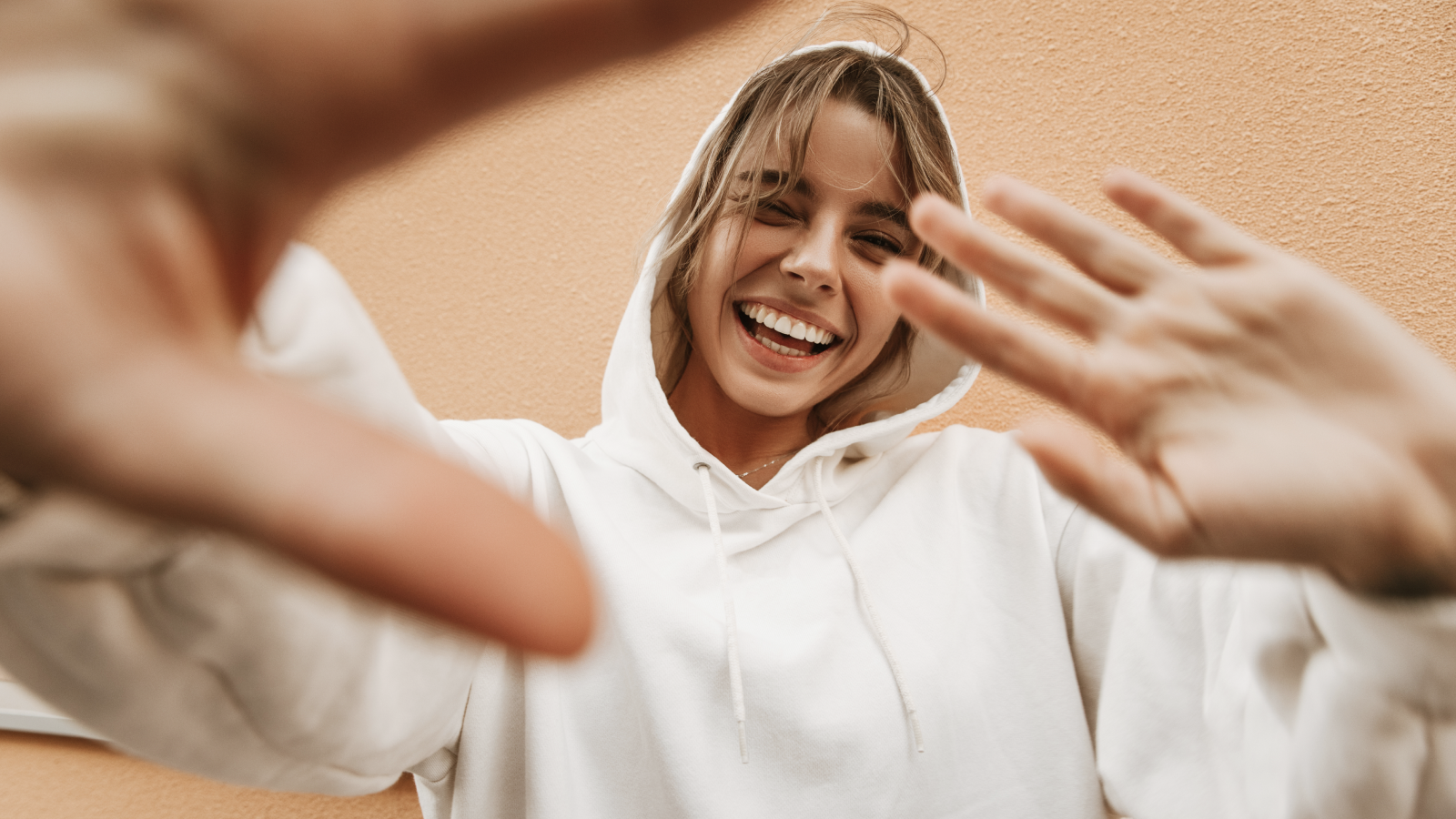 A woman in a white hoodie smiles and gestures with her hand, conveying a friendly and welcoming demeanor.