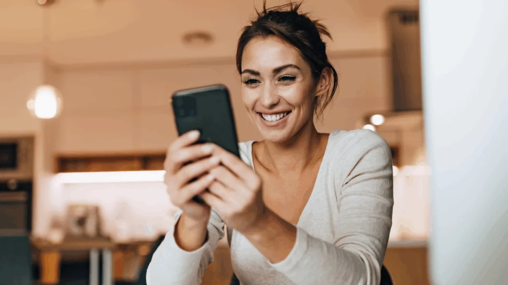 A woman smiling while using her phone, conveying a sense of joy and engagement in her activity.