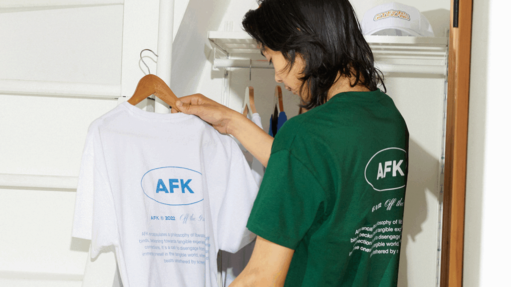A man in a green t-shirt with an “AFK” back print is holding the same t-shirt in white.