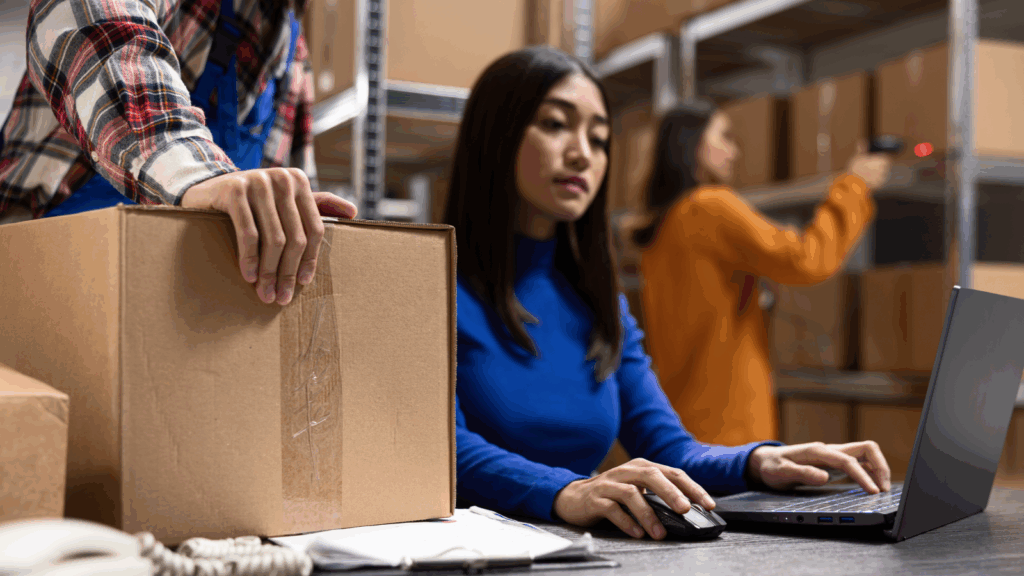 A woman focused on her laptop while seated in a busy warehouse environment.