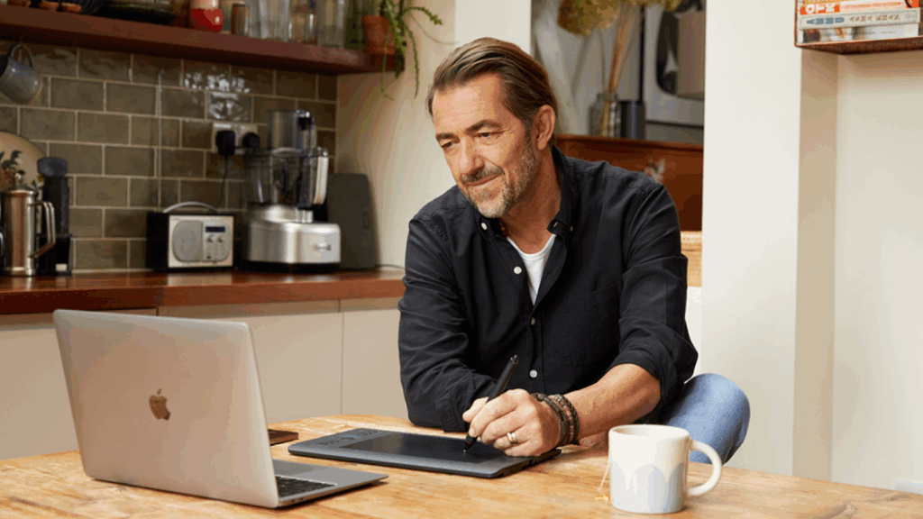 A man seated at a table, working on a laptop with a pen in hand, focused on his task.