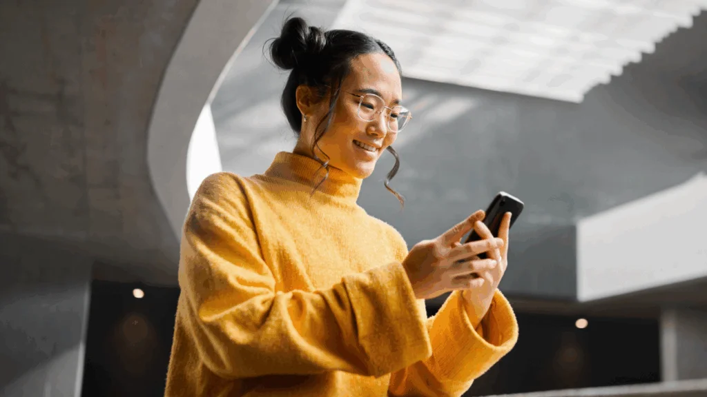 A woman in a yellow sweater gazes at her phone, appearing focused and engaged with the screen.