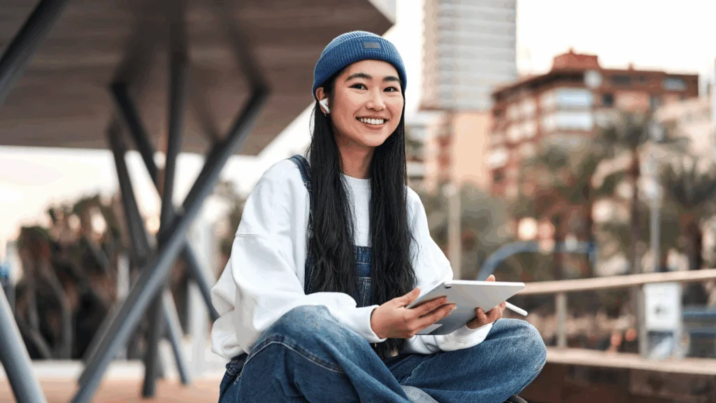 A smiling young woman is sitting in a park with a tablet.