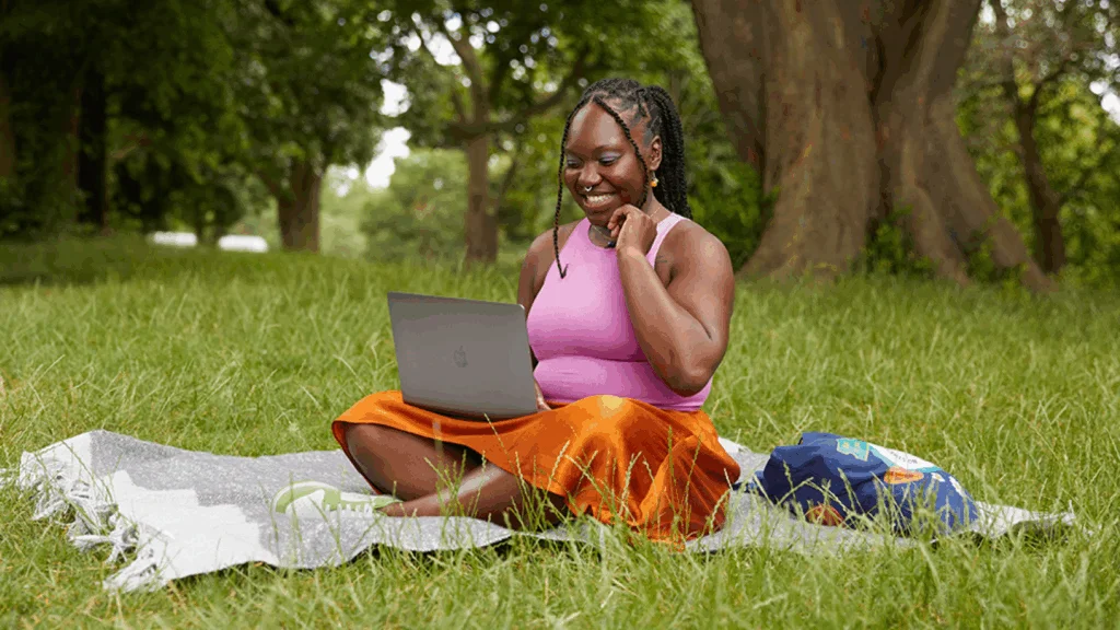 A woman sitting on the grass, focused on her laptop, surrounded by nature.