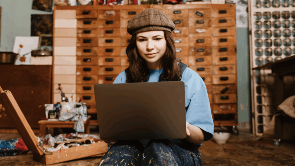 A woman artist is using a laptop while sitting on the floor in an art studio.