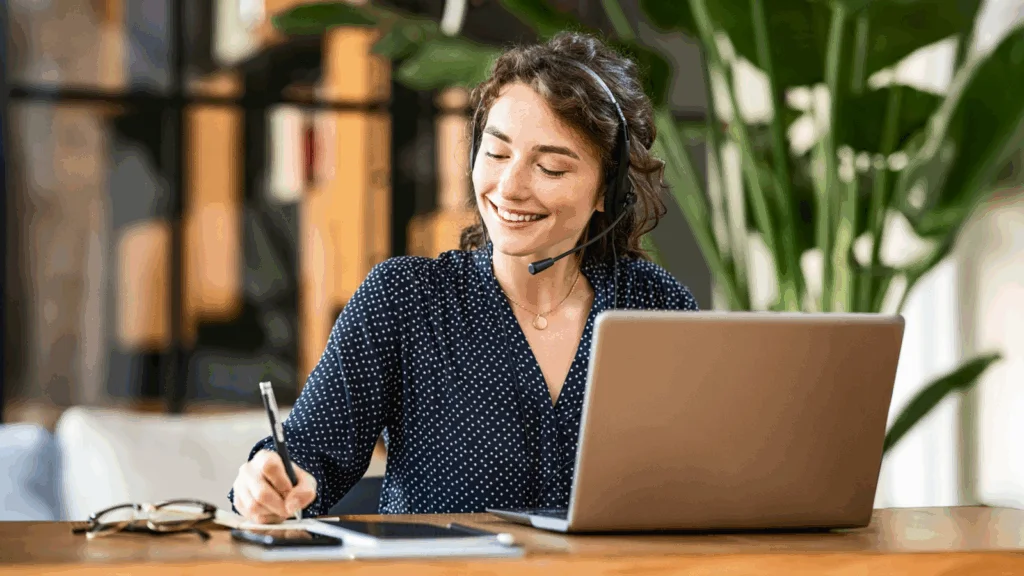 A woman wearing a headset sits at a table, working on her laptop with a focused expression.