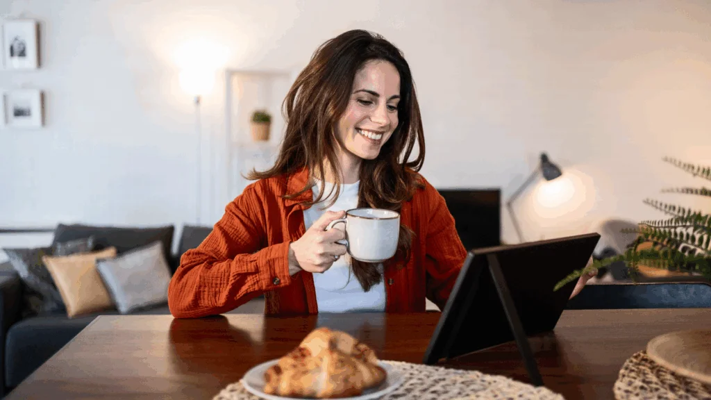 Prepare for the official holiday shopping season A smiling woman is sitting at a table, drinking coffee and using a tablet to find out when Black Friday is.
