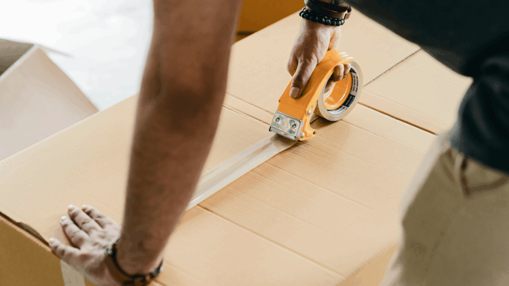 A man measures the dimensions of a box using a tape measure in a well-lit workspace.