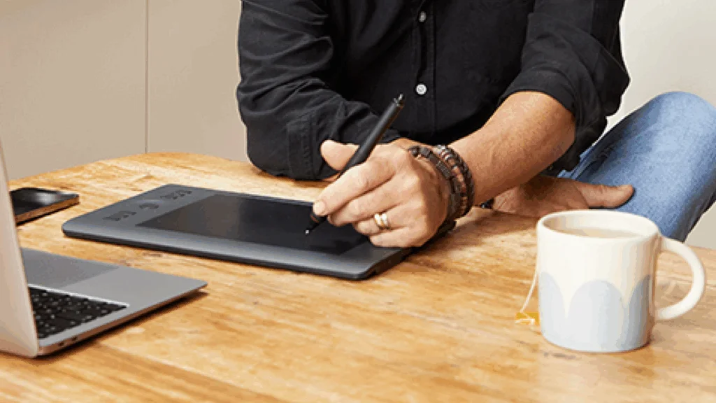 A man seated at a table, working on a laptop with a pen in hand, focused on his task.
