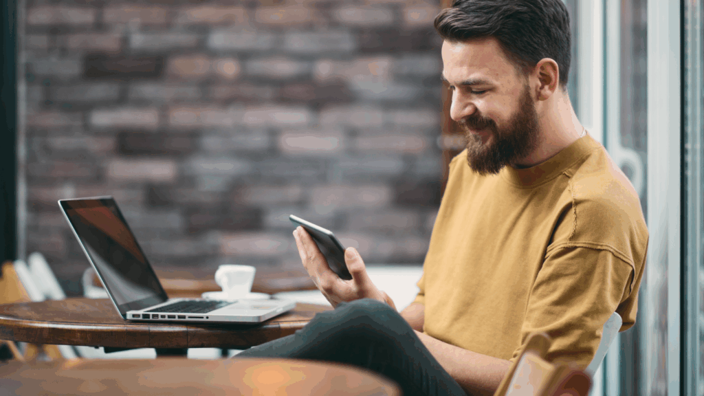 A man is looking at his mobile phone and smiling while sitting at a table with a laptop.