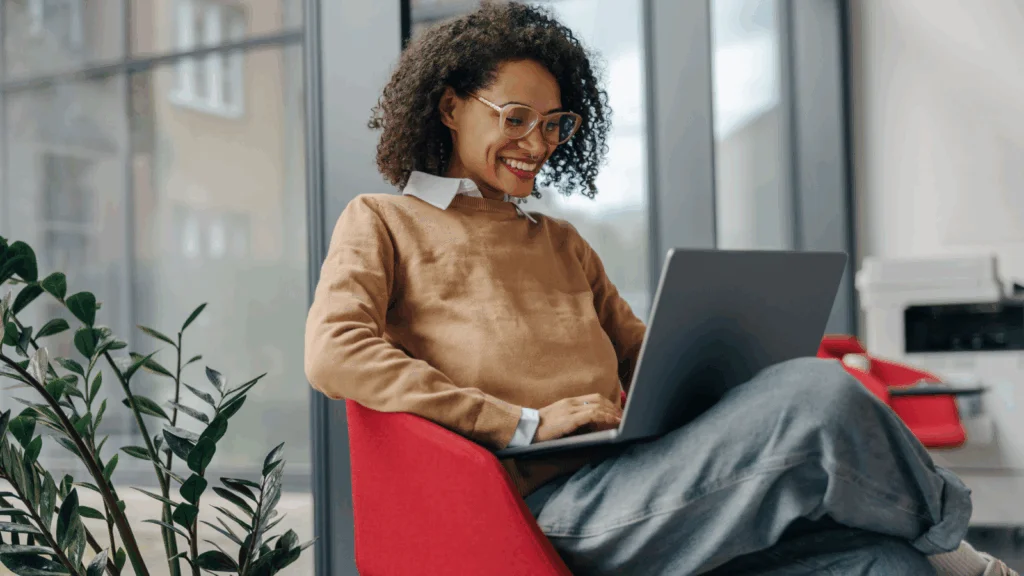 A woman seated on a chair, working on a laptop with a focused expression.