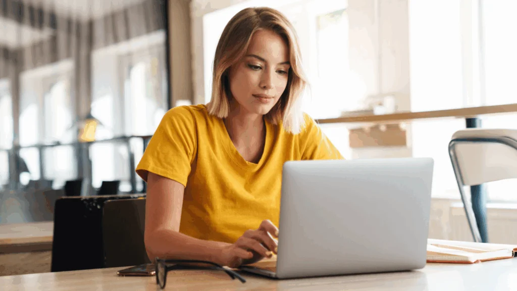 A woman in a yellow shirt sits at a table, working on a laptop.