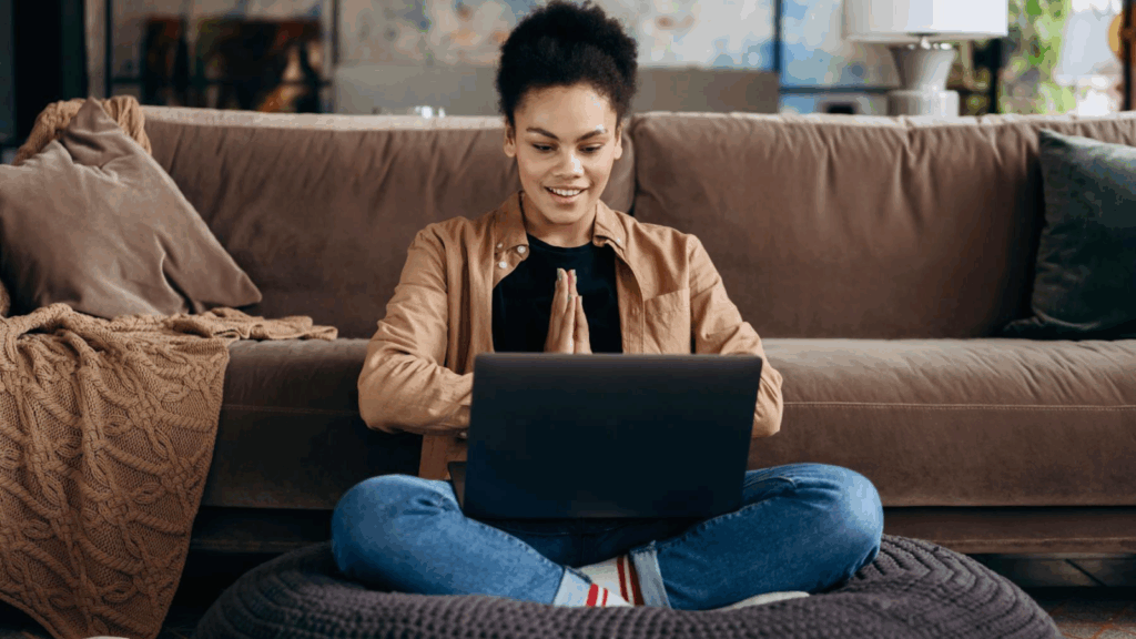 A woman sitting on a couch, focused on her laptop, with a cozy living room setting in the background.
