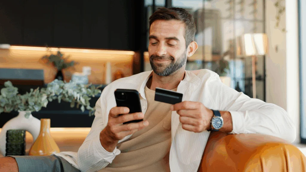 What to expect from Black Friday and what is Cyber Week A smiling man is sitting on a couch, holding a credit card and making online purchases using a mobile phone.