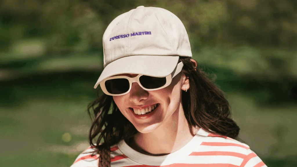 A woman with sunglasses and a hat smiles warmly at the camera, exuding a cheerful and relaxed vibe.