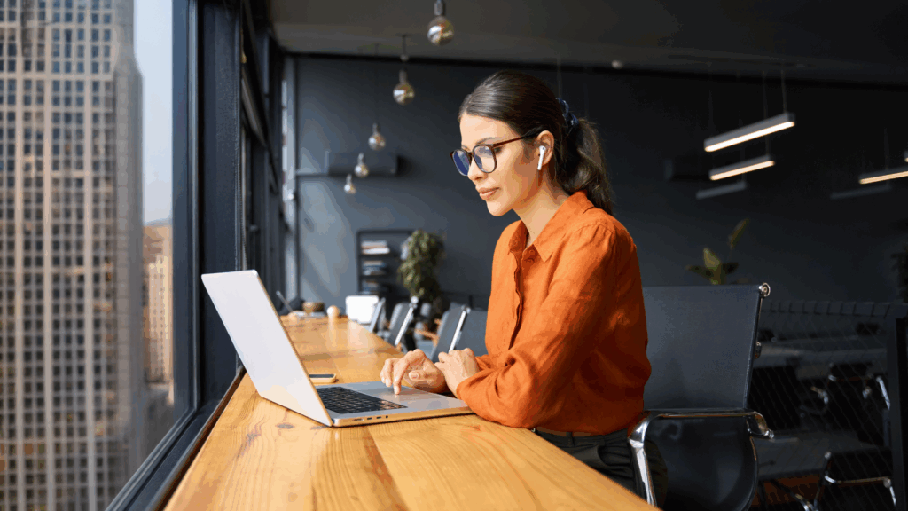 A woman in an orange shirt sits at a table, working on a laptop.