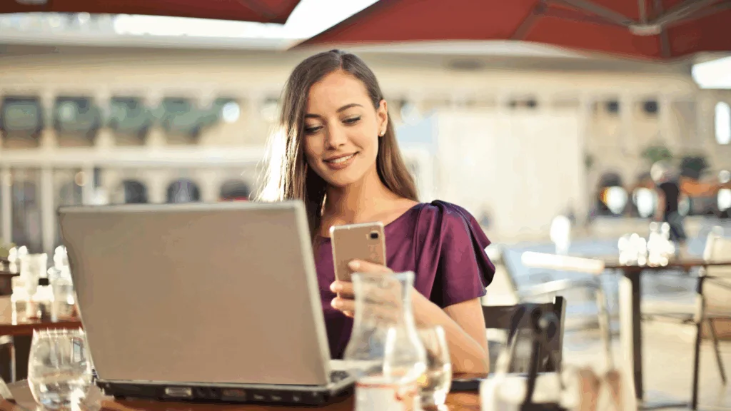 A woman seated at a table, working on a laptop with a cell phone beside her.