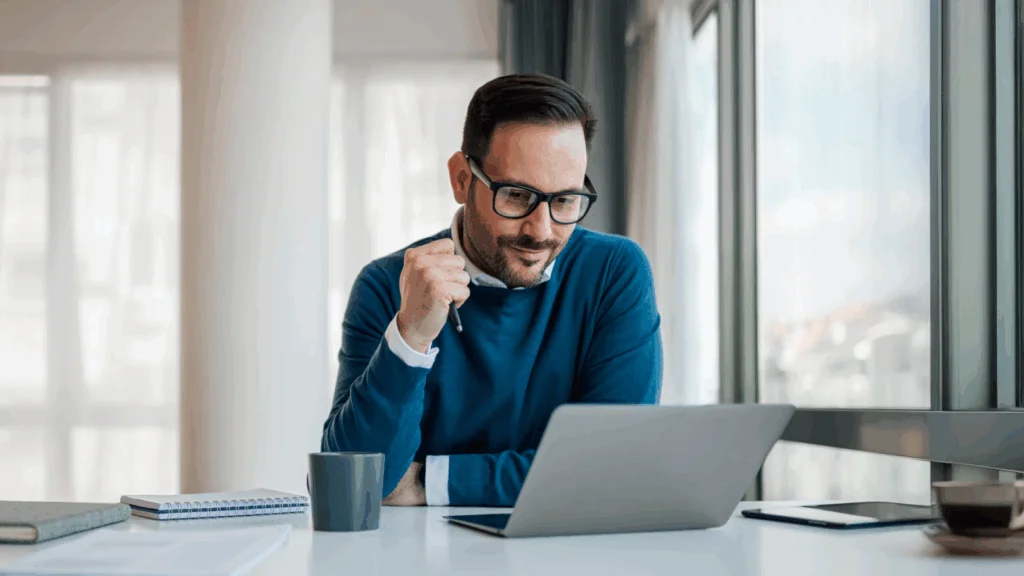 A man wearing glasses is seated at a desk, working on a laptop.