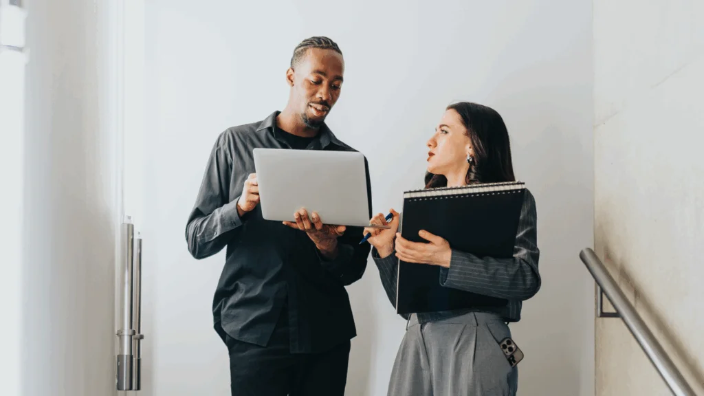 Two business professionals stand on a stairway, holding a laptop and discussing their work.