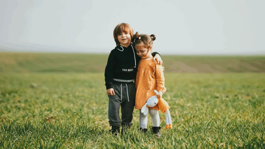 Two children standing together in a green field, smiling and enjoying a sunny day outdoors.