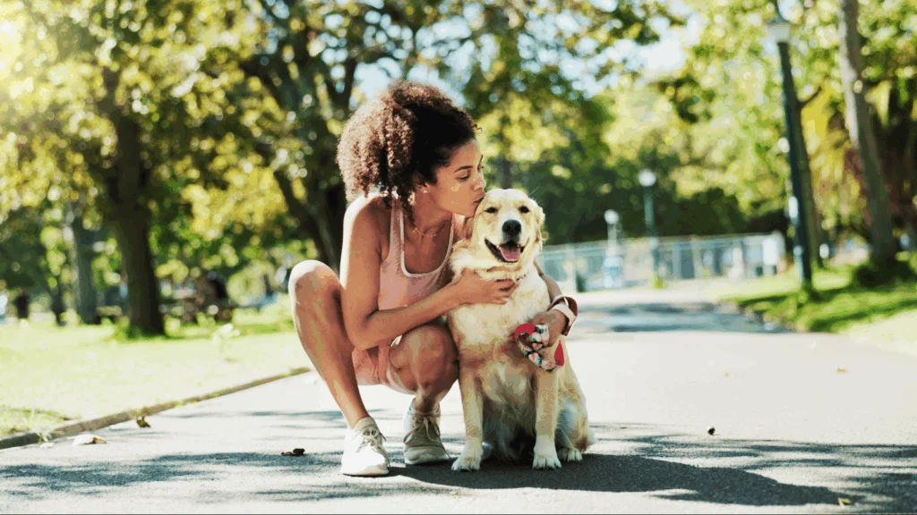 A woman kneels beside her dog in a park, enjoying a moment of companionship amidst the greenery.