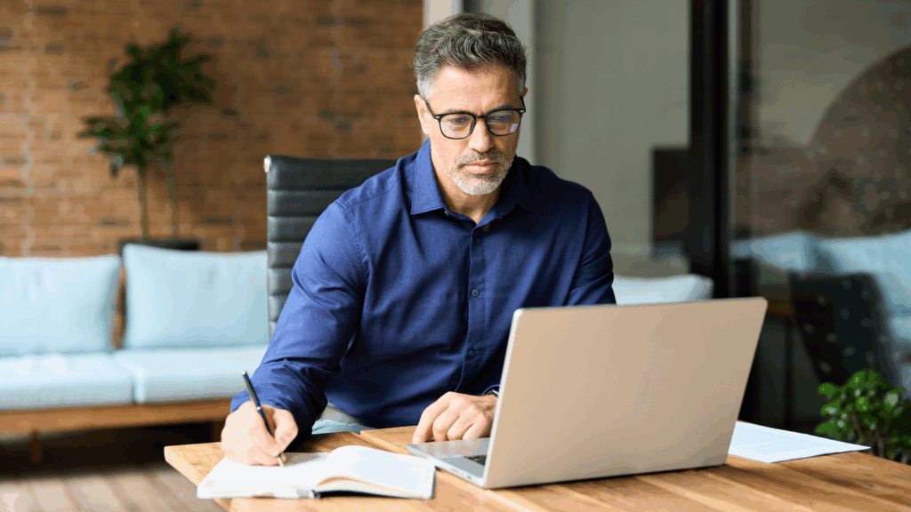 A man wearing glasses is focused on working on his laptop at a desk.