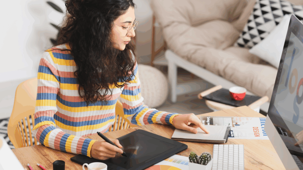 A woman sits at a desk, focused on her laptop, with a pen in hand, ready to take notes or write.