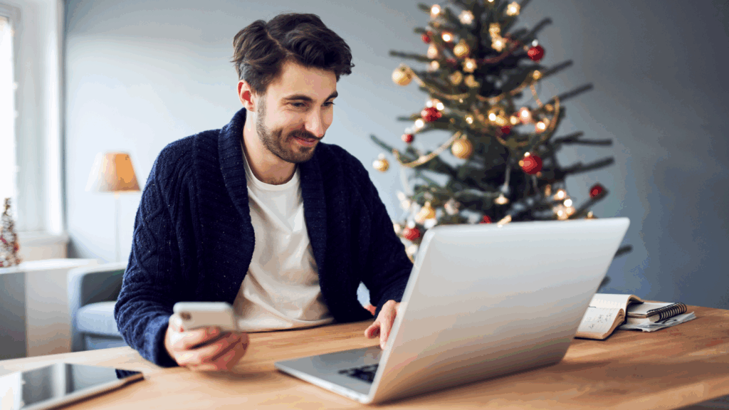 A man is holding a mobile phone and using a laptop with a Christmas tree standing behind him.