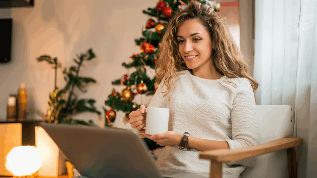 A woman sits in front of a decorated Christmas tree, using a laptop and enjoying the festive atmosphere.