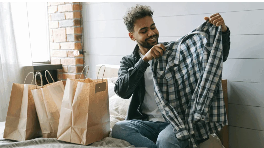 A man sits on a bed surrounded by shopping bags, appearing relaxed and content after a shopping trip.
