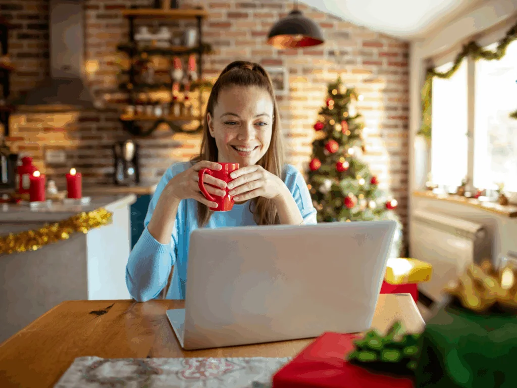 A smiling woman is sitting at a kitchen table, drinking coffee, looking at a laptop with Christmas decorations behind her.