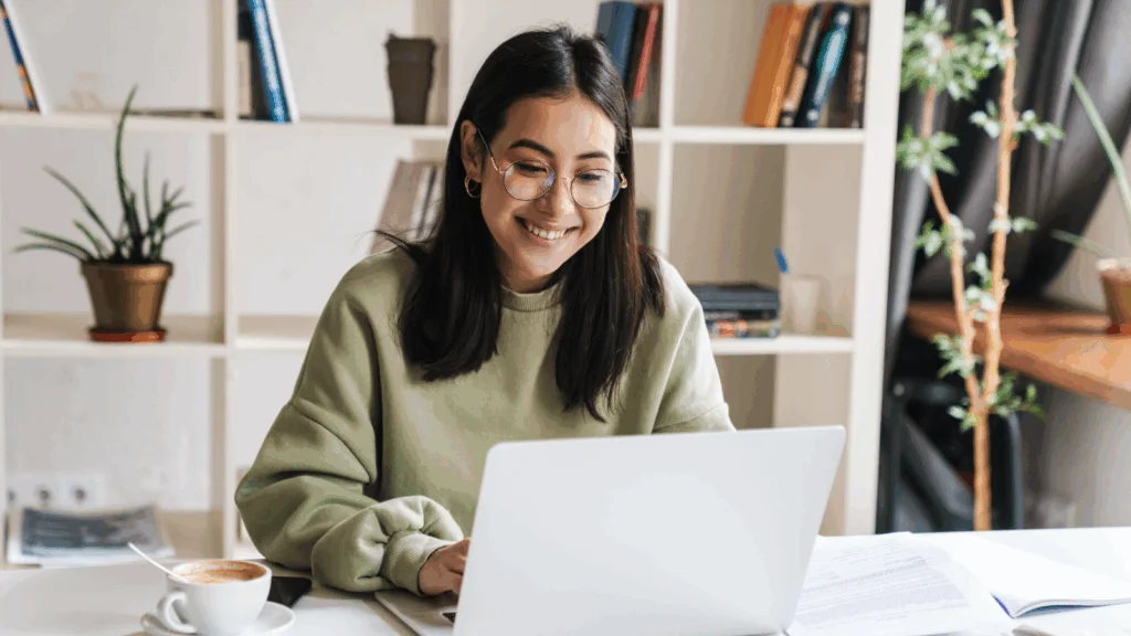 A woman smiles while working on her laptop, conveying a sense of joy and engagement in her task.