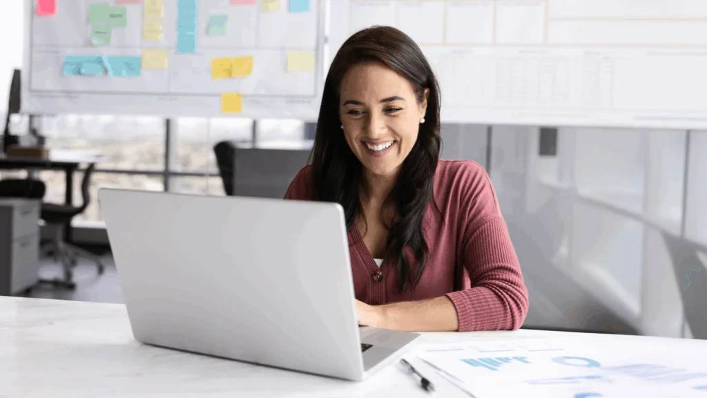 A woman smiles while working on her laptop, conveying a sense of joy and engagement in her task.