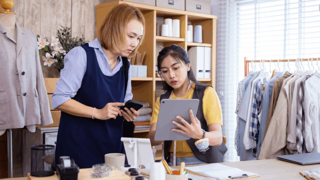 Two women in a clothing store examining a tablet together, discussing fashion options and styles.