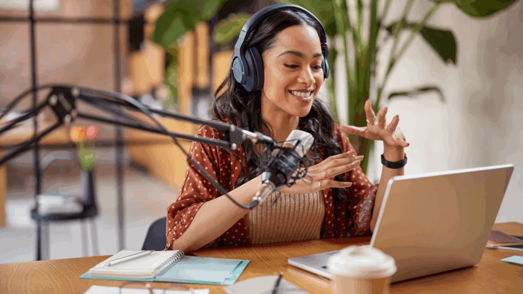A woman wearing headphones sits at a table with a laptop and microphone, focused on her work.